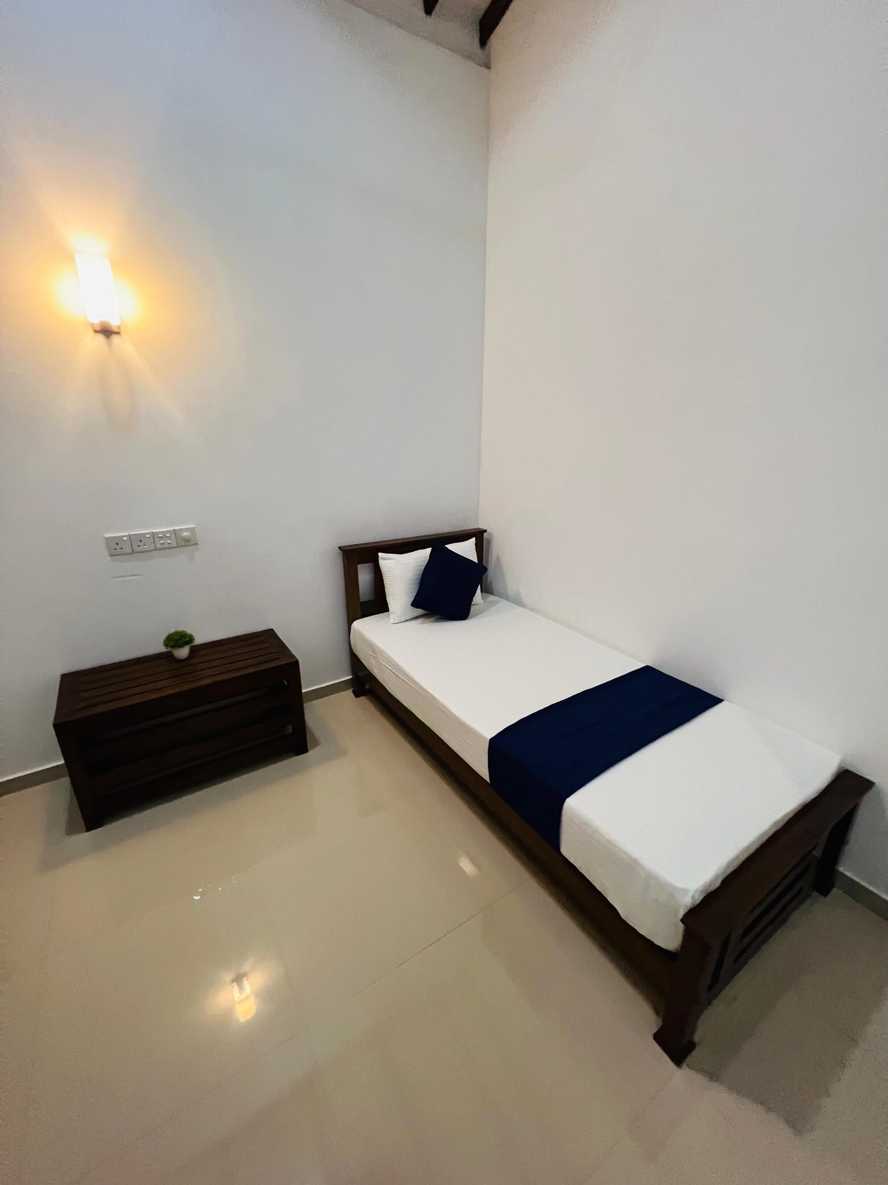 A small, neatly arranged bedroom with a single bed, a bedside table, a wall-mounted light, and a power outlet.