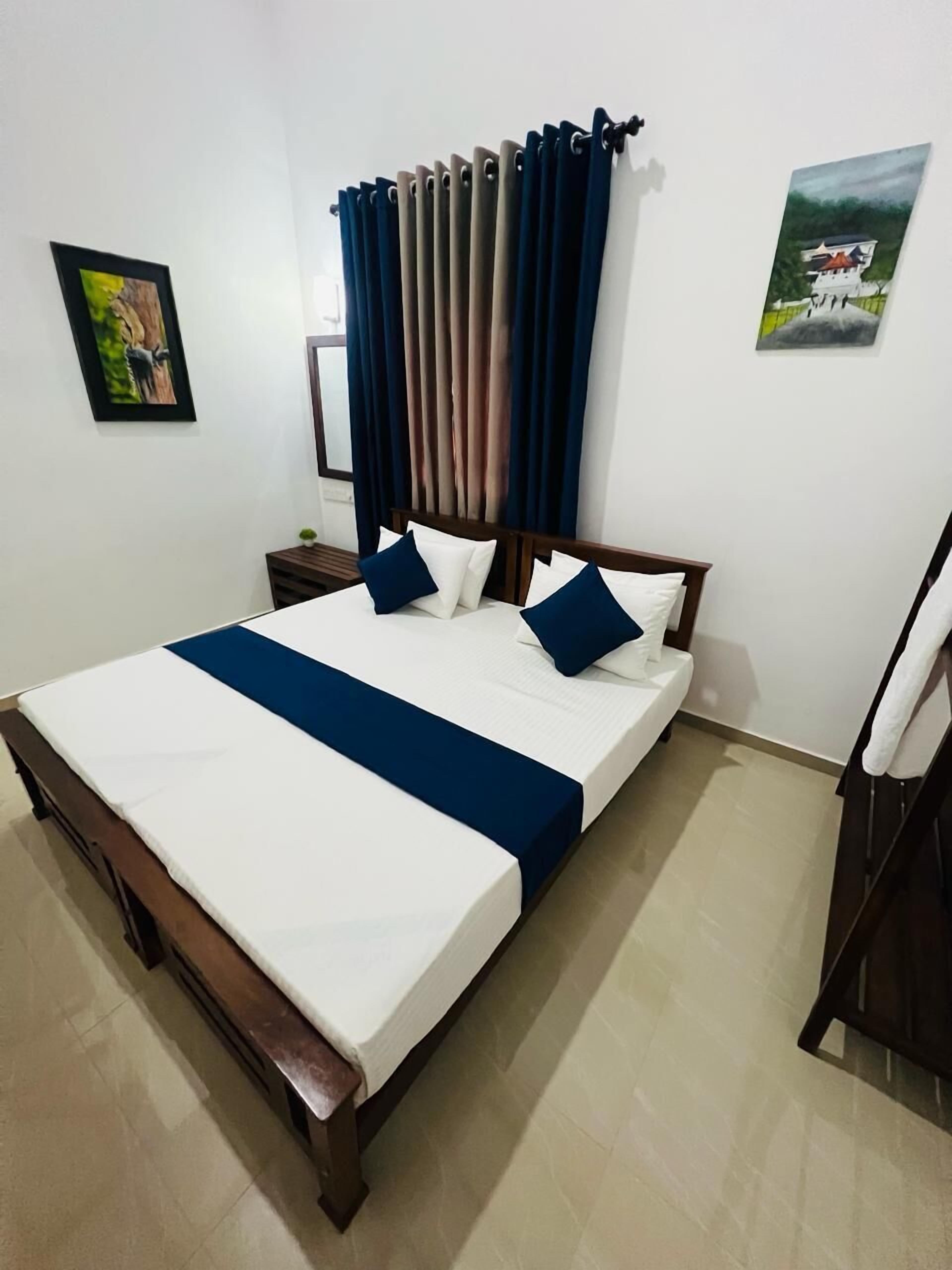 A neatly made bed with a blue bedspread and pillows, a wooden headboard, a nightstand with a lamp, and a painting on the wall.
