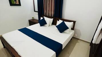 A neatly made bed with a blue bedspread and pillows, a wooden headboard, a nightstand with a lamp, and a painting on the wall.