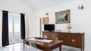 Een kamer met een houten eettafel, transparante stoelen, een televisie, een kast met een lamp en twee ingelijste foto's aan de muur.