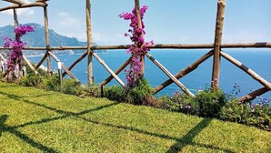Une pergola en bois, ornée de fleurs violettes, offrant une vue sur la côte et les montagnes en arrière-plan.