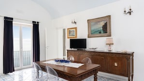 Une pièce comprenant une table à manger en bois, des chaises transparentes, un téléviseur, un meuble avec une lampe, et deux tableaux encadrés accrochés au mur.