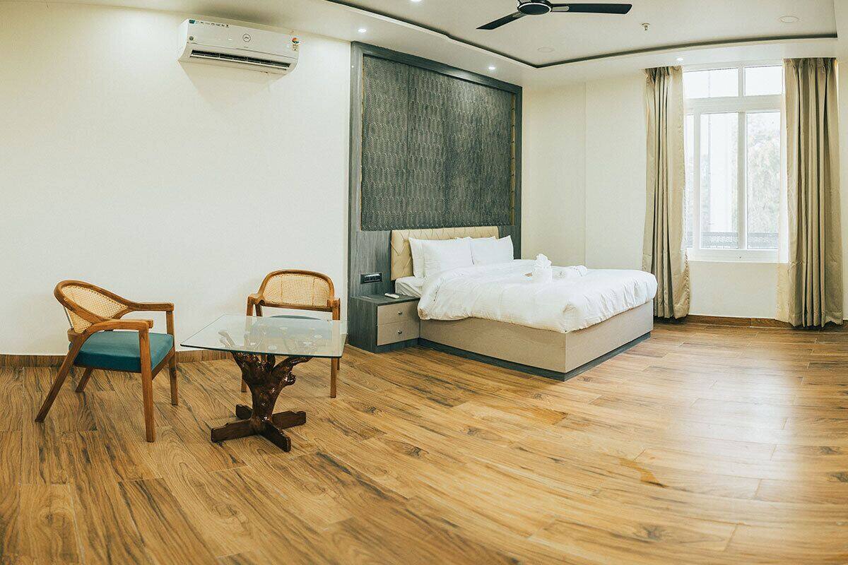 A hotel room with a bed, a glass table, two chairs, an air conditioning unit, a ceiling fan, and a window with curtains.