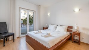 Ett hotellrum med en säng i trä, två sängbord, en stol och en balkong med gardiner.