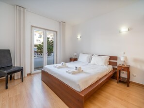 Ein Hotelzimmer mit einem hölzernen Bett, zwei Nachttischen, einem Sessel und einem Balkon mit Vorhängen.