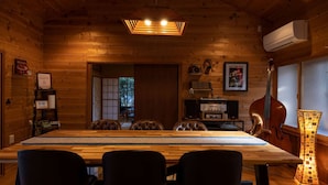 Interior de una cabaña de madera con una larga mesa de comedor, cuatro sillas, una lámpara en el techo, un aire acondicionado de pared y un cartel enmarcado.