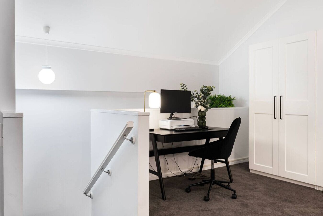 Un pequeño espacio de oficina con un escritorio, computadora y silla. Hay un armario blanco con una planta encima y una puerta blanca con manija.