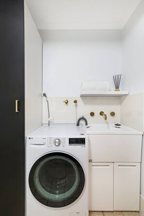 A compact laundry room with a front-loading washing machine, a sink with a faucet, and a small shelf above the sink. - Adelaide `Hills-View` Retreat (Adelaide)