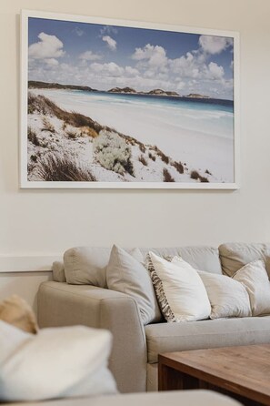 A living room with a beige sofa, a wooden coffee table, and a framed landscape painting of a beach. - The Retreat on Chaffey (Mildura)