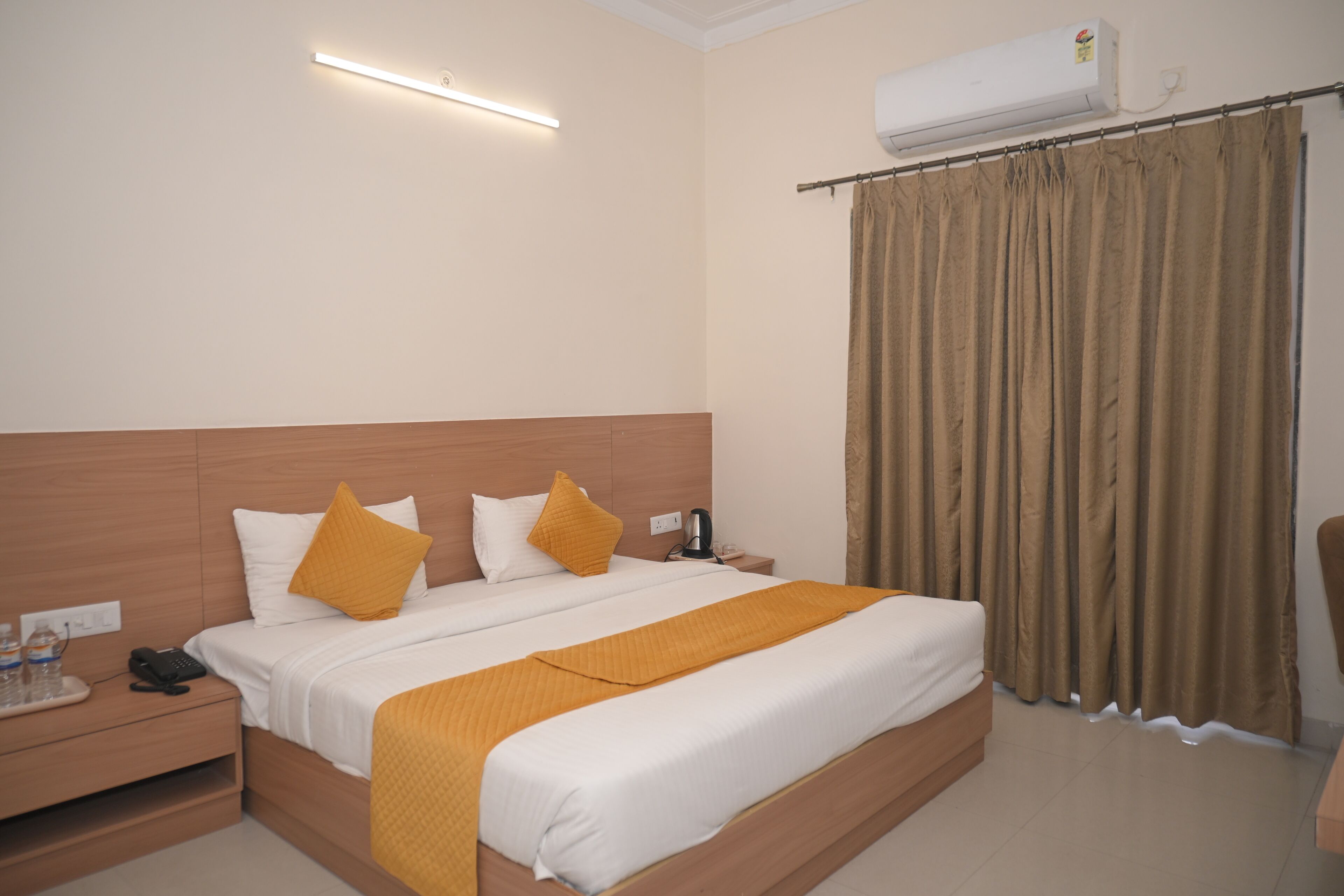 A hotel room with a large bed, a bedside table with a phone and water bottles, a nightstand, and a wall-mounted air conditioner.