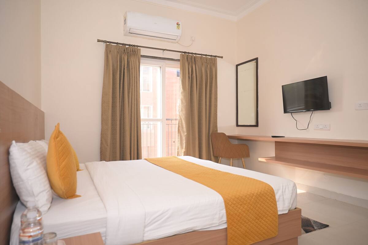 A hotel room with a bed, a TV, a desk, and a window with curtains.
