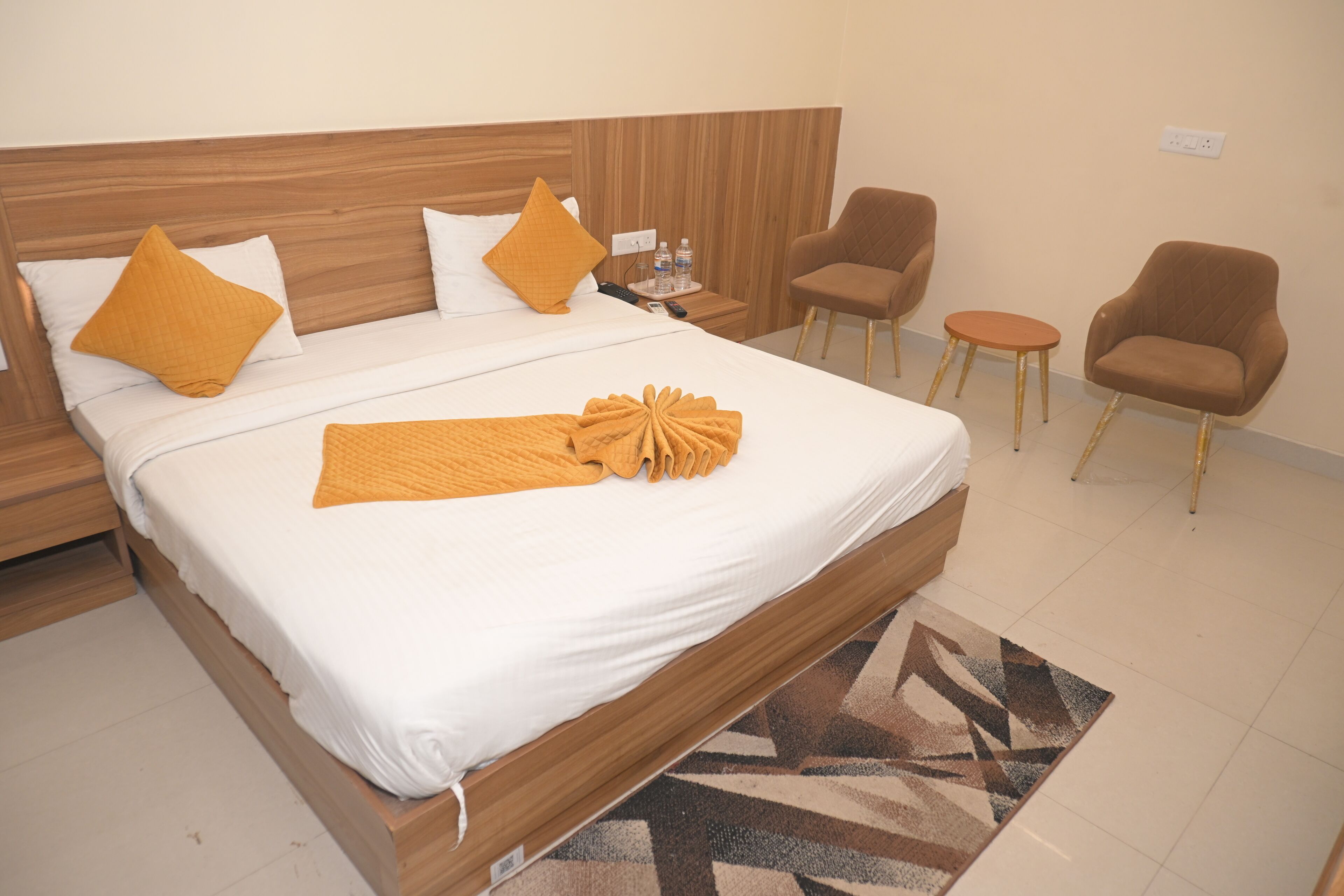 Hotelkamer met een houten bed, wit bedgoed, twee oranje kussens, een opgevouwen handdoek, een kleine tafel met waterflesjes en twee fauteuils.