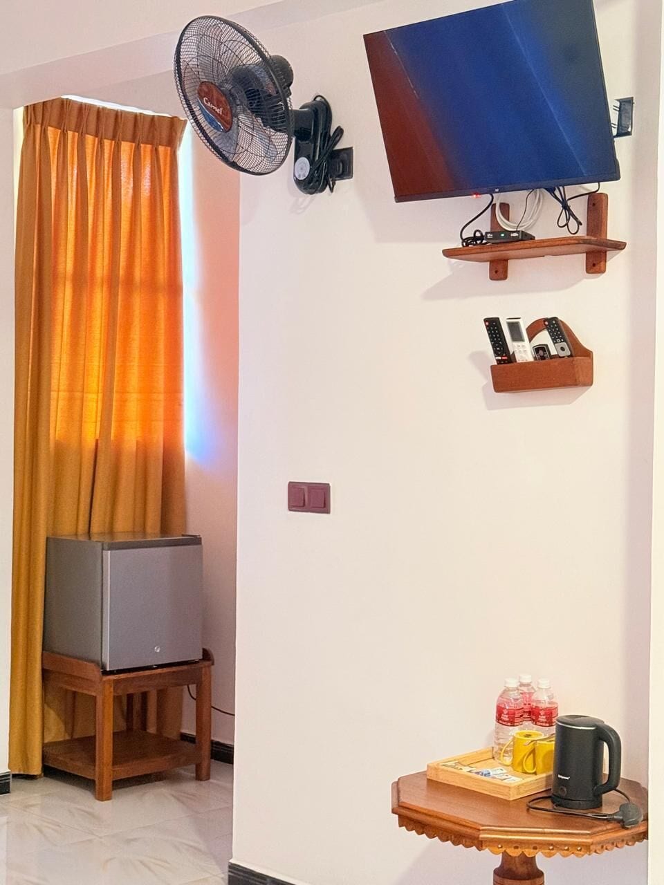 A room with a television mounted on the wall, a standing fan, a small table with a kettle and cups, and a cabinet with remote controls.