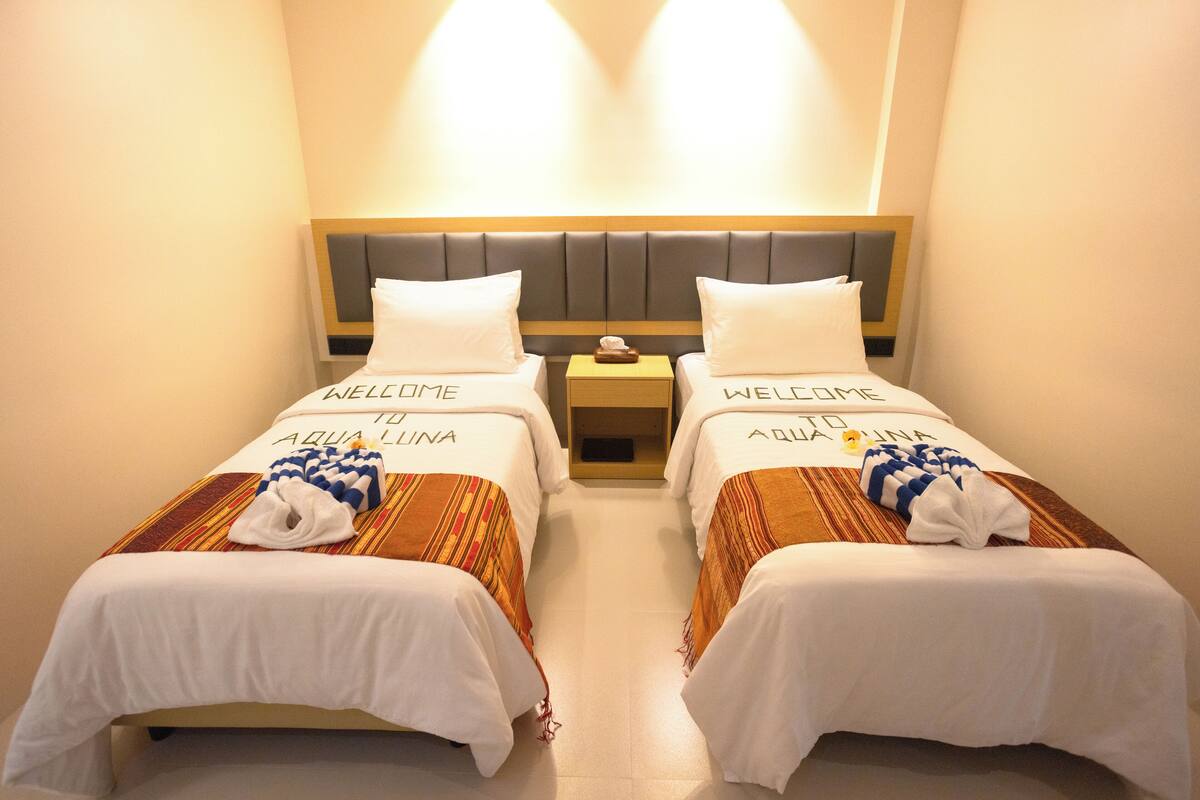 Two twin beds with "WELCOME AQUA LINA" linens, a wooden nightstand, and a wall-mounted light fixture.