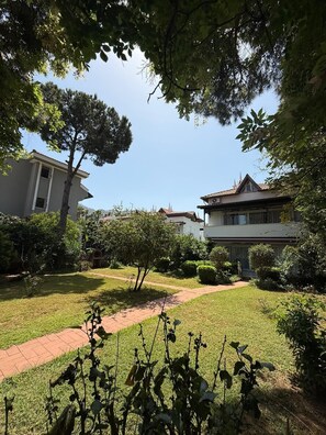 A courtyard with a pathway, surrounded by greenery and two buildings with distinct architectural styles. - Mete 3 Villa İçmeler Weekly Daily Rentals (Marmaris)