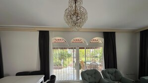 A spacious room with a dining table, two upholstered chairs, a chandelier, and a view through a glass door to an outdoor patio area. - Mete 3 Villa İçmeler Weekly Daily Rentals (Marmaris)