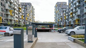 Parkkialue, jossa on useita autoja, jalankulkijoiden suojatie sekä parvekkeilla varustettu rakennus ja katettu sisäänkäynti.