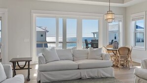 Un salón amplio con un sofá blanco grande, una mesa de comedor con sillas y vista al océano a través de ventanas amplias.
