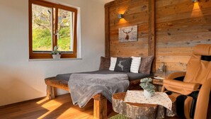 Een gezellige kamer met een bed, een houten bankje, een leren fauteuil, een tafeltje met een plant en een raam met uitzicht op groen.