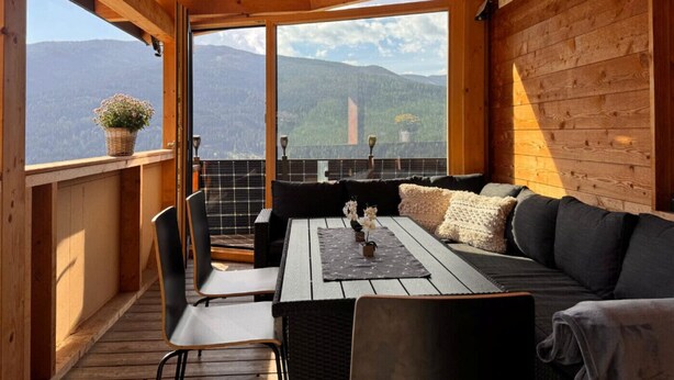 Een houten balkon met een bank, stoelen en een tafel, met uitzicht op een bergachtig landschap.