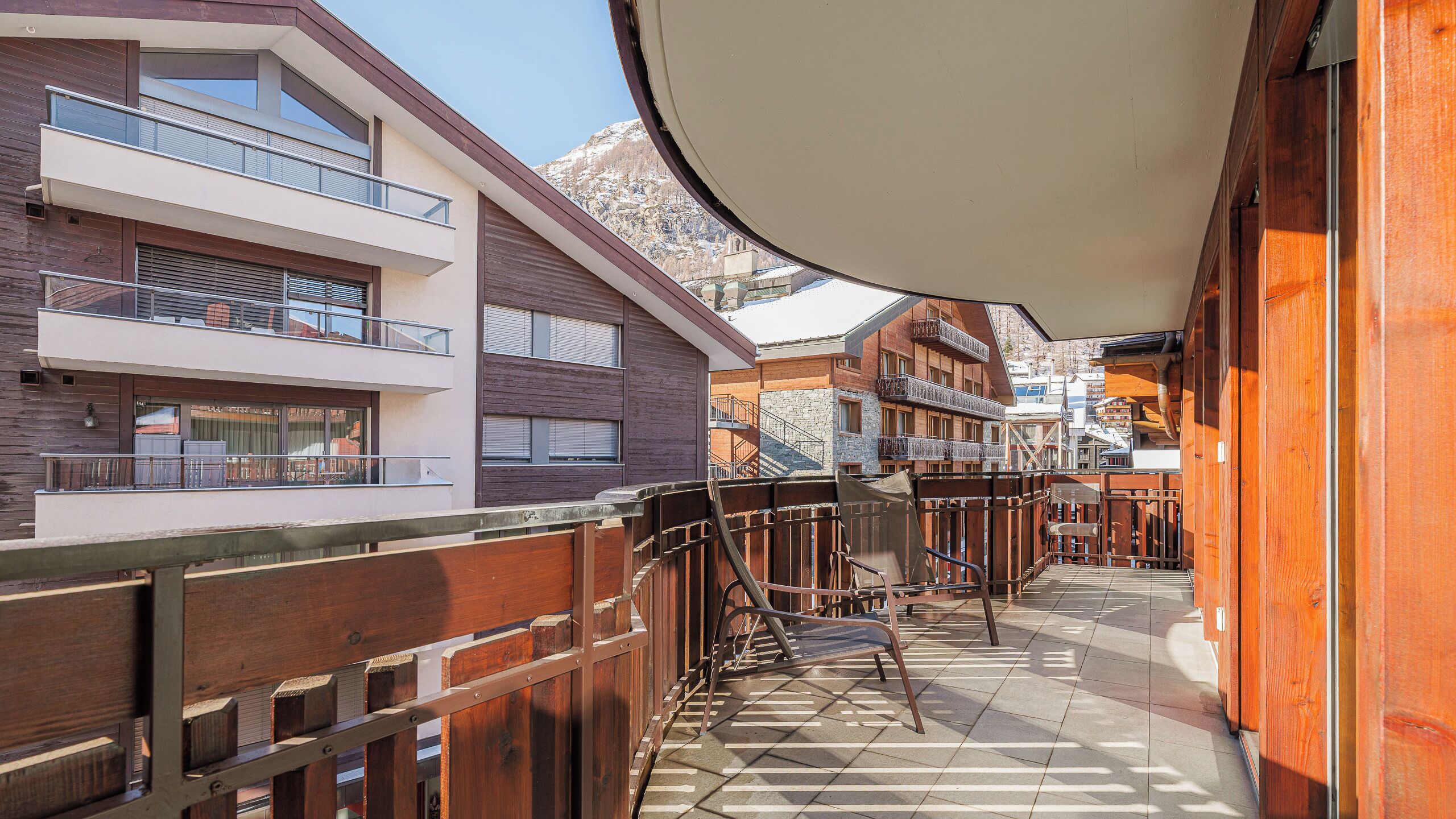 Un corridoio con ringhiere in legno, pavimento piastrellato e vista su edifici con balconi e finestre.