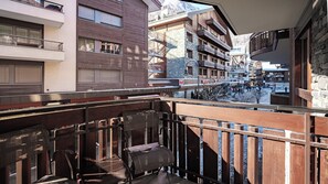 A balcony with a view of a town square, featuring multi-story buildings with balconies, a sign for 'MIGROS', and a wooden fence. - Haus Zur Matte by Zermatt Premium (Zermatt)