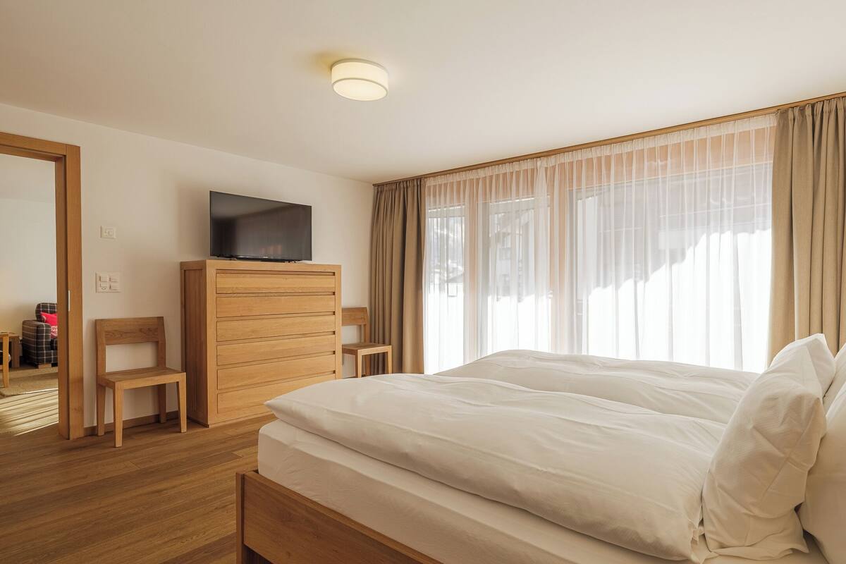 A hotel room with a large bed, wooden dresser, television, and a view of the outdoors through sheer curtains.
