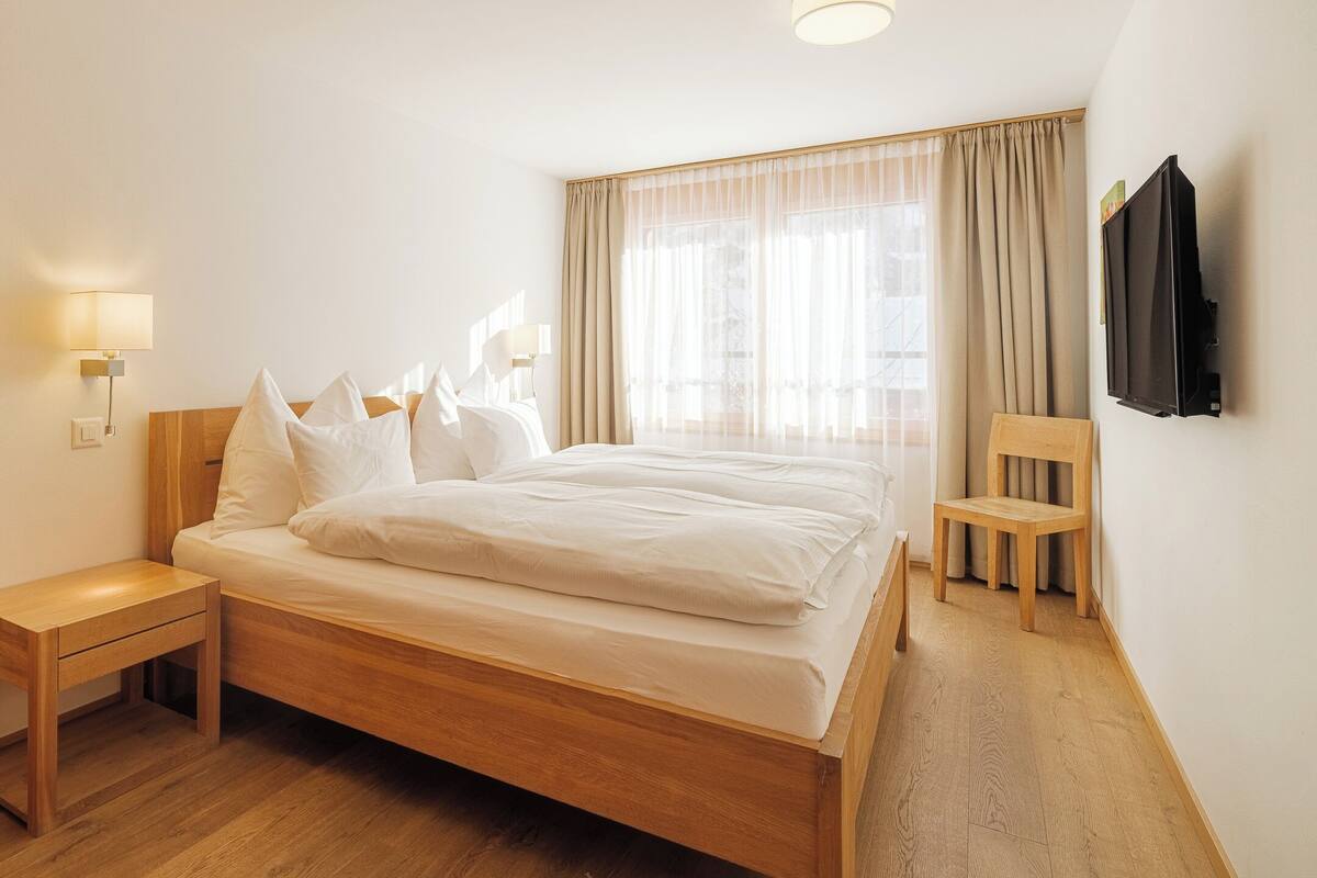 A hotel room with a wooden bed, a nightstand, a chair, a TV mounted on the wall, and a window with curtains.