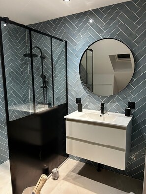 A modern bathroom with a herringbone tile backsplash, a round mirror, a white vanity with a black faucet, and a glass-enclosed shower area. - To the hillside butterfly (Magenta)