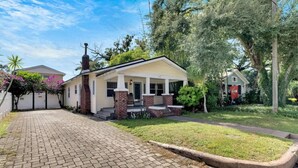 Uma casa térrea com chaminé de tijolos, varanda frontal com degraus e entrada por um caminho pavimentado.