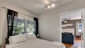 Quarto com cama, janela com persianas, ventilador de teto e vista para uma cozinha.