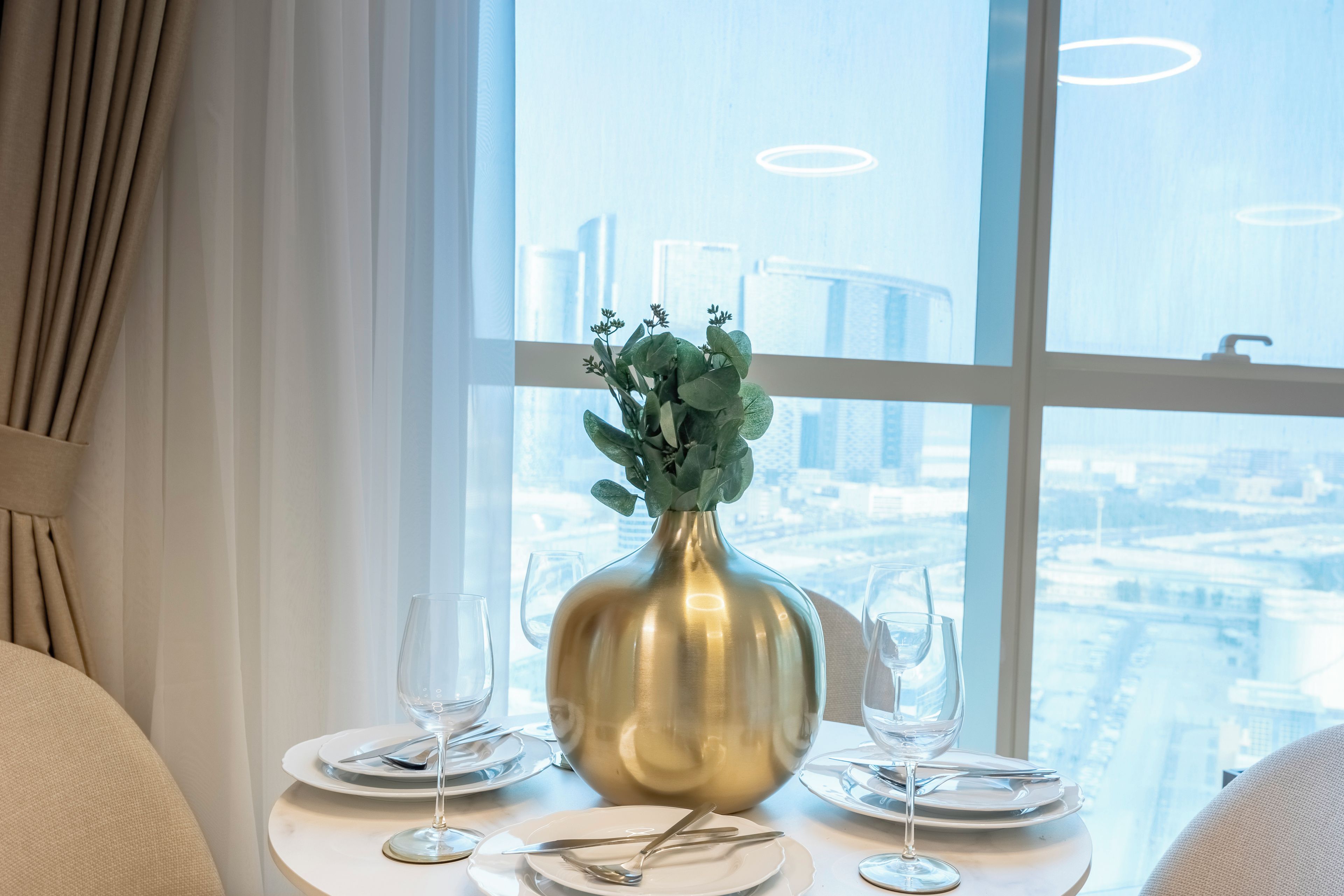Een eettafel gedekt voor twee personen, met een vaas groen, twee wijnglazen en bestek, geplaatst bij een raam met uitzicht op een stadsbeeld.