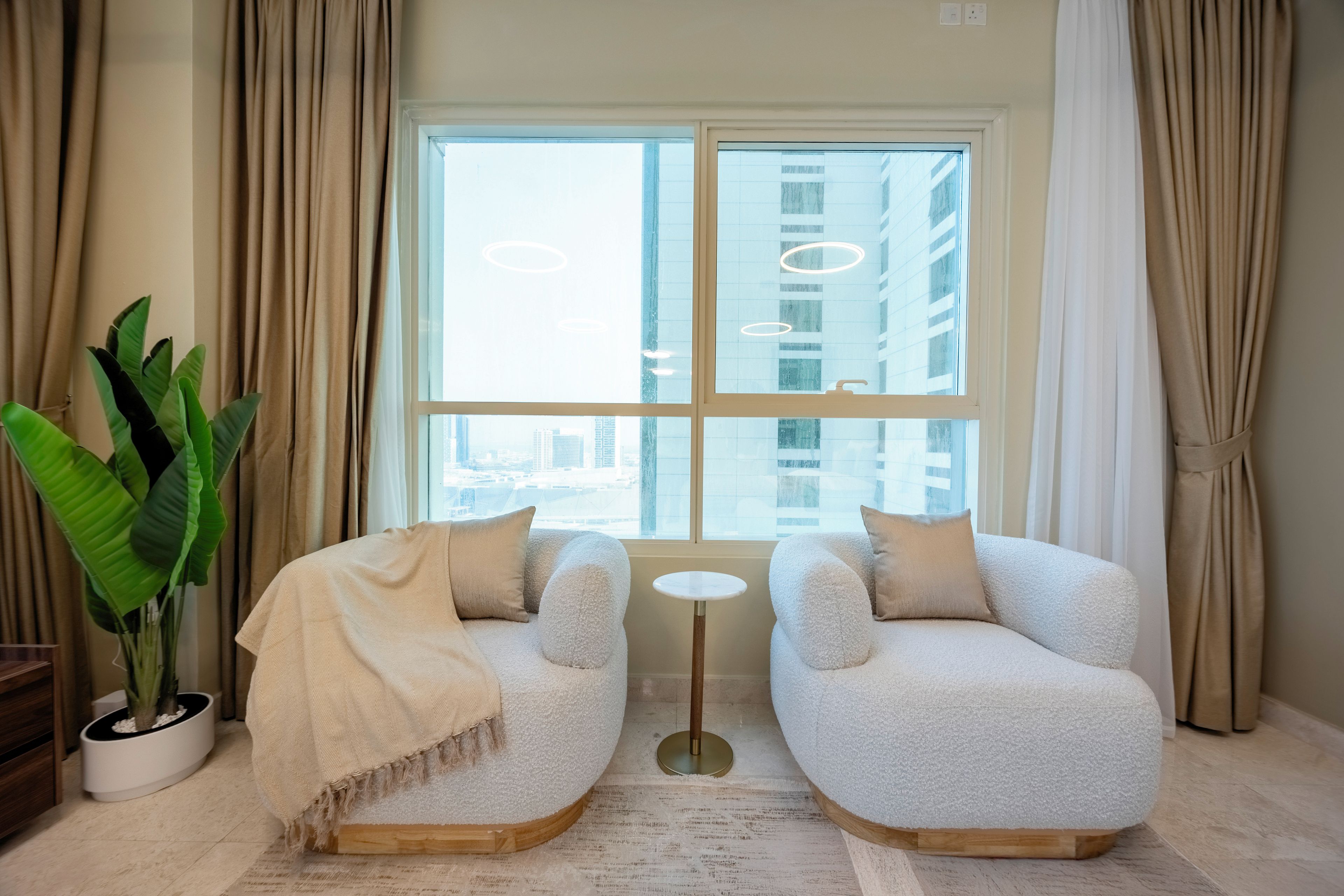 Een moderne kamer met twee comfortabele fauteuils, een klein rond tafeltje, een groot raam met uitzicht op de stad en een potplant.