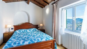 Quarto com teto de madeira, cama com colcha florida azul, duas luminárias de apoio, janela com vista para uma cidade e um radiador.