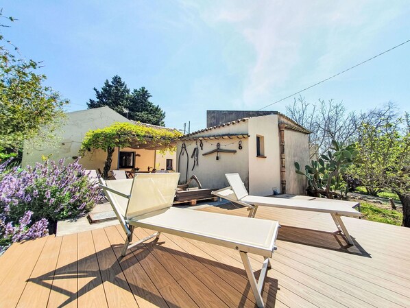 Una terraza de madera con dos sillones, un pequeño edificio con techo de tejas y un jardín con flores moradas.