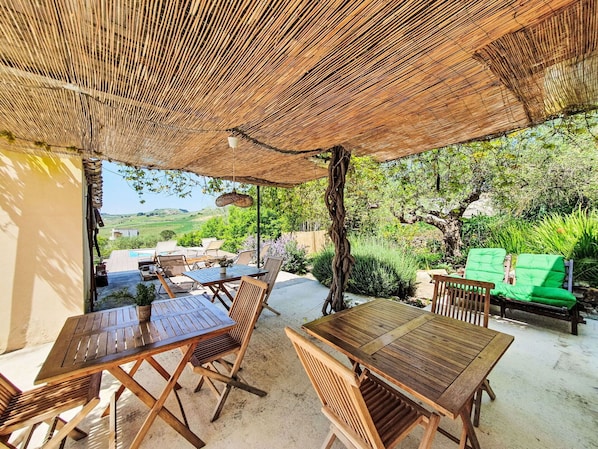 An outdoor patio with wooden tables and chairs, a woven canopy, and a view of a green landscape.