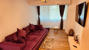 Una sala de estar moderna con un amplio sofá modular, una mesa de centro de vidrio, una mesita blanca y un televisor montado en la pared.
