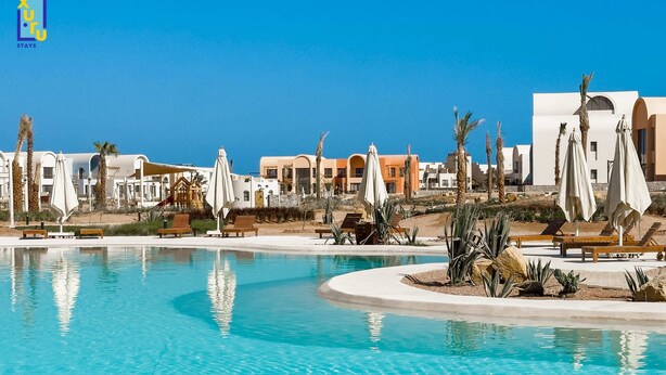 Ein Poolbereich eines Resorts mit mehreren Liegestühlen und Sonnenschirmen, umgeben von Palmen und modernen Gebäuden.