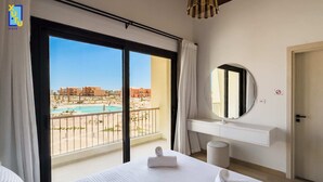 Ein Hotelzimmer mit großem Fenster und Blick auf einen Pool sowie Gebäude. Ein Bett mit weißen Bettwäsche und ein runder Spiegel an der Wand sind zu sehen.