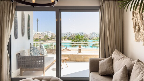 A modern living room with a sofa, a round ottoman, and a view of a pool and buildings through large windows. - Genial 2bdr @ Swan Lake El Gouna (El Gouna)