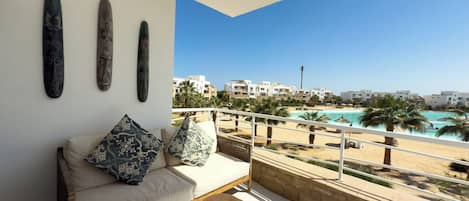 A balcony with a sofa, pillows, and a small wooden stool. It overlooks a beach with a pool and buildings in the background.