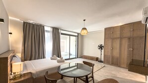 Una habitación de hotel moderna con una cama grande, una mesa de vidrio, dos sillas, un escritorio con una lámpara, un armario y un ventanal con cortinas.