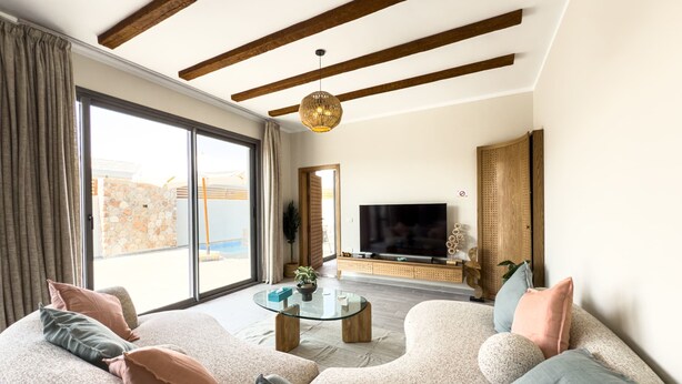 Una sala de estar moderna con un sofá grande, una mesa de centro de vidrio y un televisor. Hay un ventanal con cortinas, un techo de madera y vistas al exterior.
