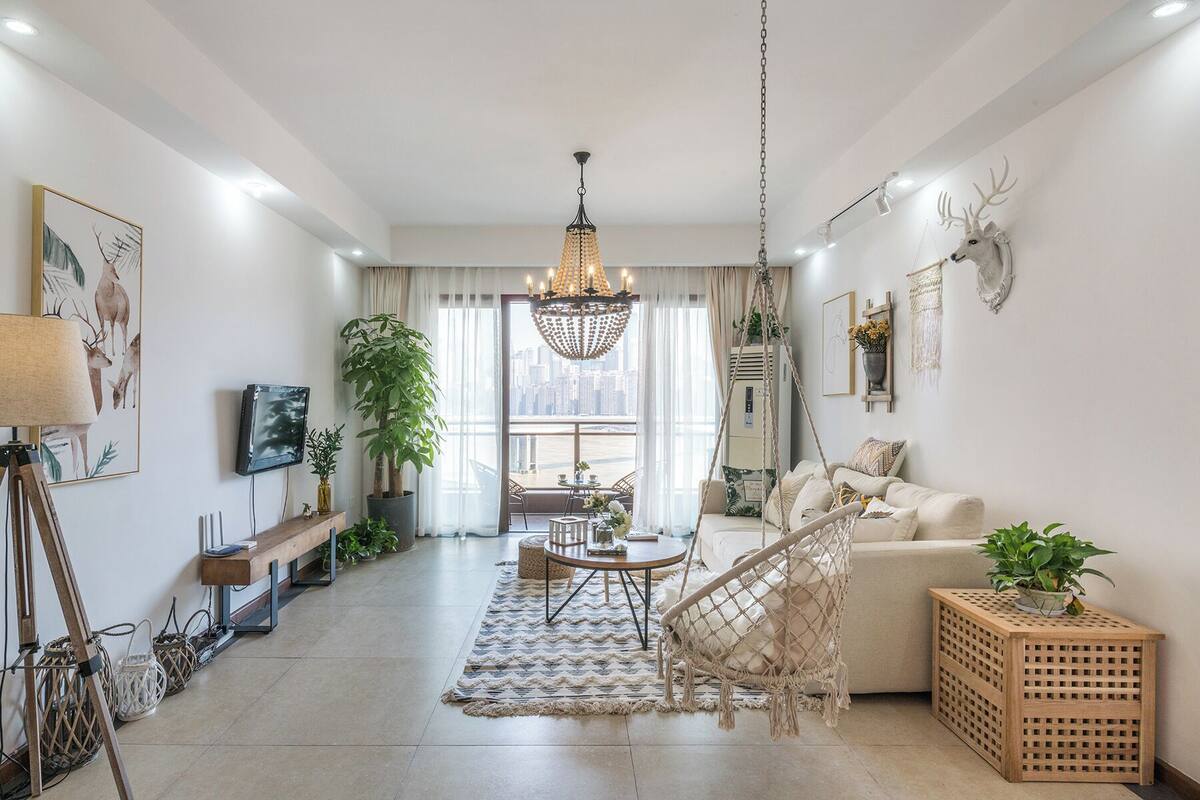 A modern living room with a sofa, a hanging chair, a chandelier, and a city view through the window.