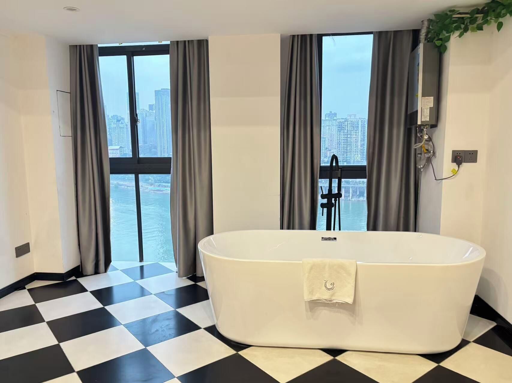 Un baño moderno con una bañera independiente, ventanas amplias con cortinas, suelo a cuadros y vistas a la ciudad.