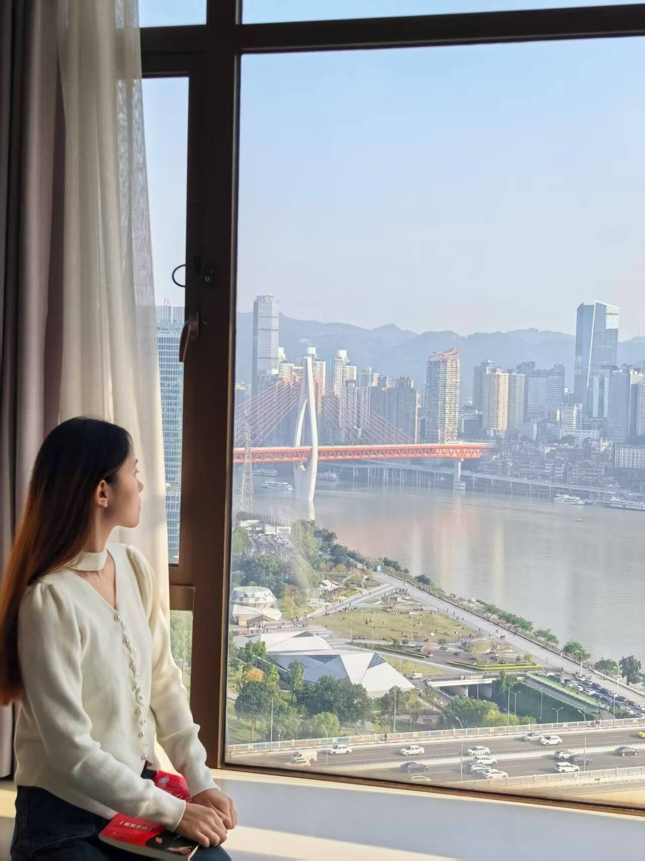 A person sitting by a window overlooking a cityscape with modern buildings and a prominent bridge.