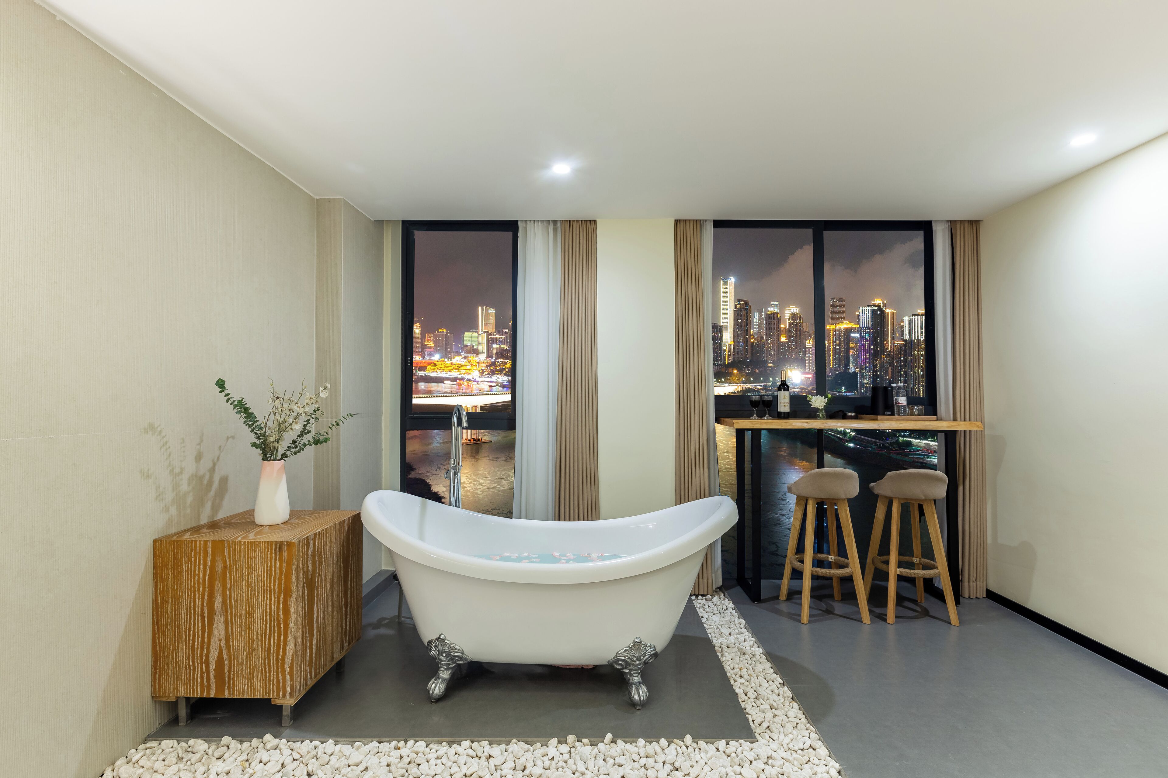 Un baño moderno con una bañera independiente, una mesita de madera con un florero y vistas a la ciudad a través de amplios ventanales.
