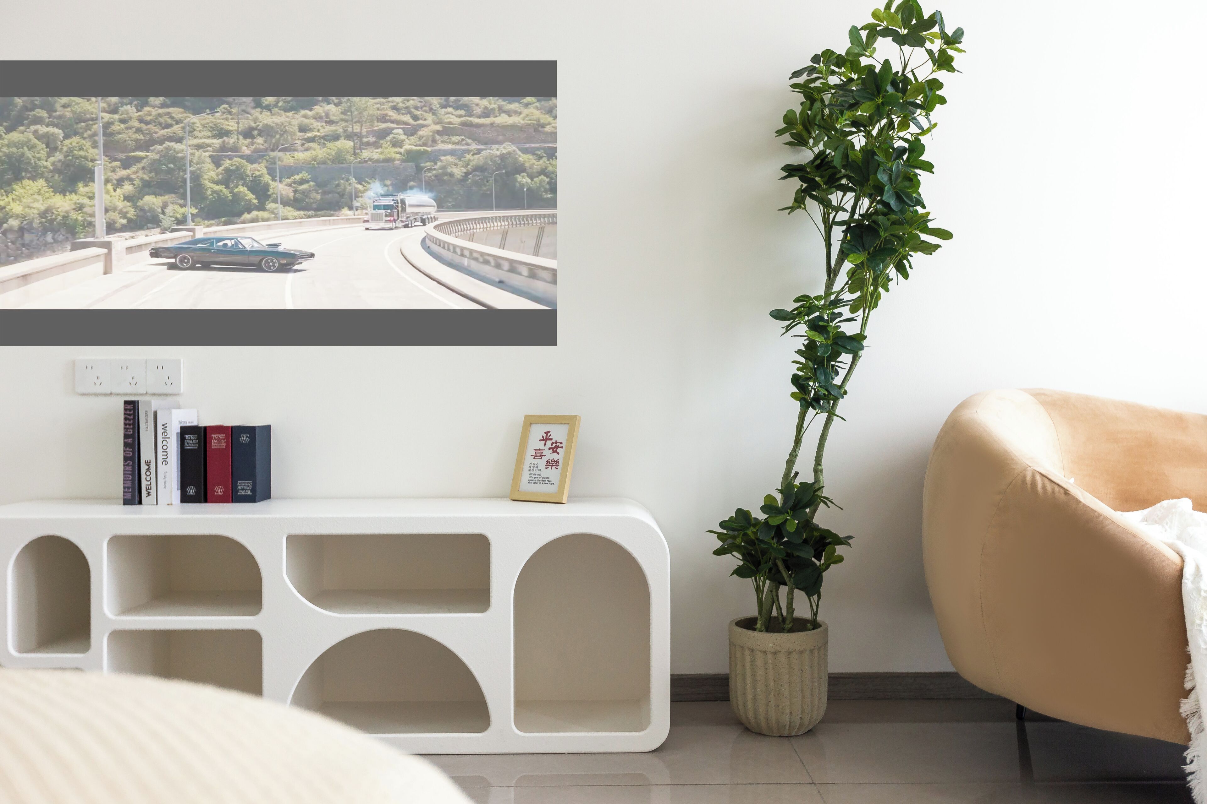 A modern living room with a white console table, a potted plant, a beige armchair, and a wall-mounted TV displaying a scenic road view.