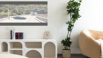 A modern living room with a white console table, a potted plant, a beige armchair, and a wall-mounted TV displaying a scenic road view.