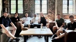 Un grupo de personas sentadas alrededor de una mesa en una habitación con paredes de ladrillo, ventanas amplias y obras de arte en las paredes.